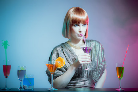 Glamorous Woman with Red Hair Wearing Shiny Retro Gown and Sipping Cocktail Drink from Straw, Standing at Bar in Disco Night Clubの写真素材