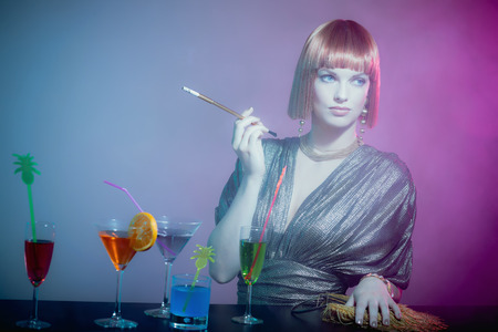 Glamorous Sophisticated Woman with Red Hair Wearing Shiny Retro Gown Smoking Cigarette at Bar Covered in Cocktails in Smoky Disco Night Clubの写真素材