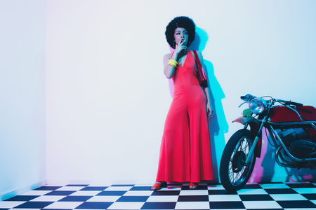 Stylish Young Lady Smoking a Cigarette Beside her Vintage Motorcycle While Looking Into Distance and...