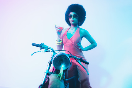 Elegant Lady with Curly Short Hair, Holding a Cocktail on her Vintage Motorcycle While Looking Into Distance Against White Background.の写真素材