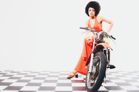 Beauty and glamour portrait of an elegant lady with an afro hairstyle posing full length on a motorbike in a long orange dress over a checkered black and white floorの写真素材
