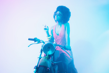 Elegant Young Woman Posing on her Vintage Motorcycle While Smoking a Cigarette Against White Background.の写真素材