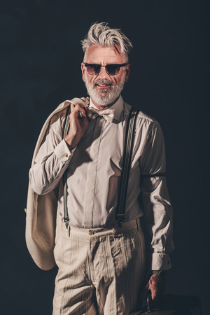 Portrait of a Stylish Bearded Senior Man with Sunglasses, Carrying his Coat and Suitcase and Smiling at the Camera Against Black Background.の写真素材