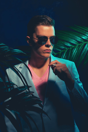 Close up Handsome Young Man Wearing Formal Suit and Sunglasses, Standing Between Palm Leaves While Holding a Cigarette and Looking Into the Distance on a Dimmer Light.の写真素材