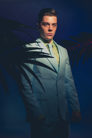 Handsome Man in Formal Wear Standing Between Palm Leaves Against Dark Blue Violet Background.の写真素材