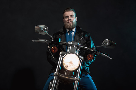 Stylish handsome bearded businessman waiting on his motorbike with a glowing headlamp looking at the camera with a serious expression, dark backgroundの写真素材