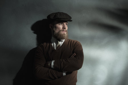 Trendy confident man in a cloth cap standing leaning against a grey wall with folded arms staring towards a beam of lightの写真素材