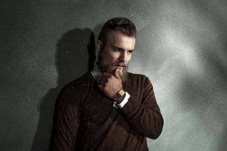 Thoughtful worried bearded young man with his hand to his chin standing staring at the floor, upper...