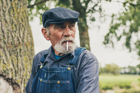 Senior grey-haired bearded country farmer standing smoking in his cloth cap and denim dungarees as he stares thoughtfully into the distanceの写真素材