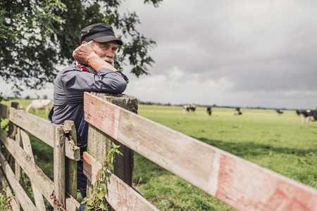 Elderly farm worker tending his cattle in the pasture standing leaning on a wooden fence with a serious contemplative expressionの写真素材