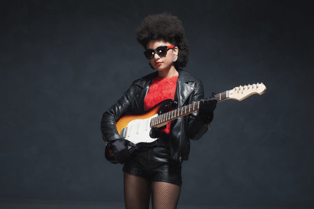 Three Quarter Shot of a Stylish Rocker Girl with Afro Hairstyle, Holding a Guitar Against Black...