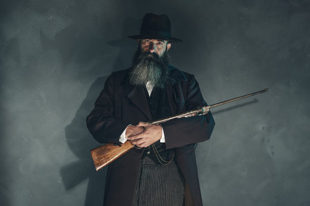 Spooky vintage beard man with rifle in 1900 style fashion against grey wall.