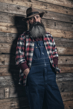 Vintage worker man with long gray beard in jeans dungarees. Standing in front of wooden wall.の写真素材