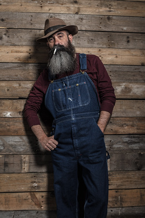 Smoking vintage worker man with long gray beard in jeans dungarees. Standing against wooden wall.の写真素材