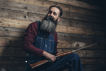 Vintage farmer holding rifle standing against wooden wall in barn.の写真素材
