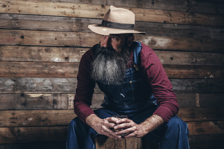 Vintage worker man with long gray beard in jeans dungarees holding whiskey. Sitting on wooden crate in barn.の写真素材