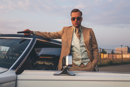Retro 1970s gangster with pistol leaning against vintage car.の写真素材