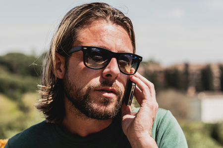 Casual man with long blonde hair and sunglasses calling with mobile phone outdoors.の写真素材