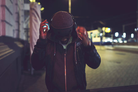 Urban afro man with woolen cap and headphones in city at night with blurred street lights.