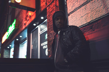 Urban fashion afro man with woolen cap and hoody standing against wall with city lights at night.の写真素材