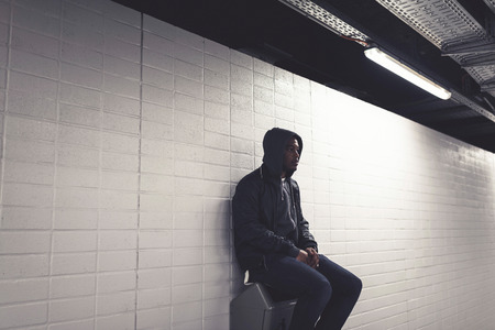 Urban fashion afro man with hoody sitting on garbage bin against white wall in subway station.の写真素材