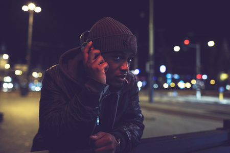 Urban afro man with woolen cap and headphones in city at night with blurred street lights.の写真素材