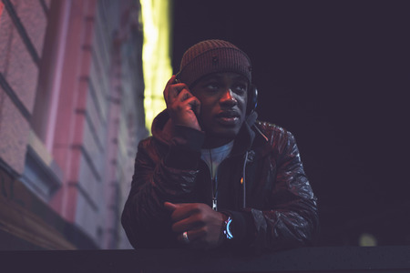 Urban afro man with woolen cap and headphones in city at night with blurred street lights.の写真素材