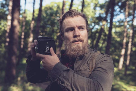 Alert beard man holding vintage camera in forest.