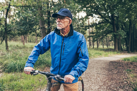 Resting senior man standing with bicycle on forest trail.の写真素材