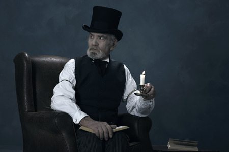 Dickens Scrooge Man Sitting in Chair with Book by Candlelight.の写真素材