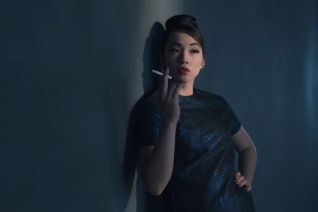 Retro 40s styled Asian woman in evening dress smoking cigarette. Leaning against wall.の写真素材