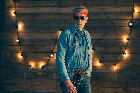 Senior bearded guitarist holding guitar standing in front of wooden wall with light bulbs.の写真素材
