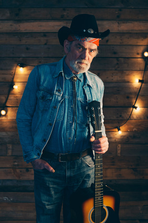 Senior bearded country and western musician holding guitar standing in front of wooden wall with light bulbs.の写真素材