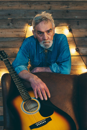 Senior musician with beard leaning on back of chair with acoustic guitar.