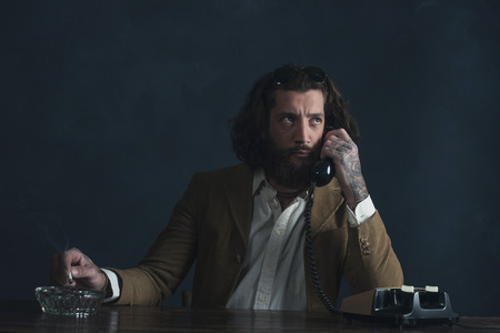 Retro 1970 undercover agent with beard calling behind desk while smoking cigarette.の写真素材