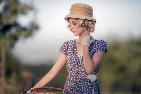 Vintage 1930s fashion woman in blue summer dress standing with bicycle in rural landscape.の写真素材
