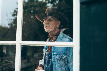Farmer with beard and hat holding cup of coffee looking out window of old farm.の写真素材