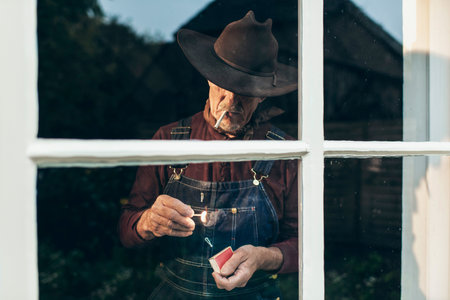 Senior man with beard and hat lighting cigarette while standing behind window.の写真素材
