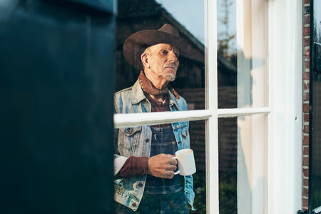 Farmer with beard and hat holding cup of coffee looking out window of old farm.の写真素材