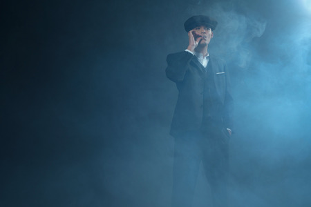 Retro 1920s english gangster wearing suit and flat cap standing in smoky room. Smoking cigarette. Peaky blinders style.の写真素材