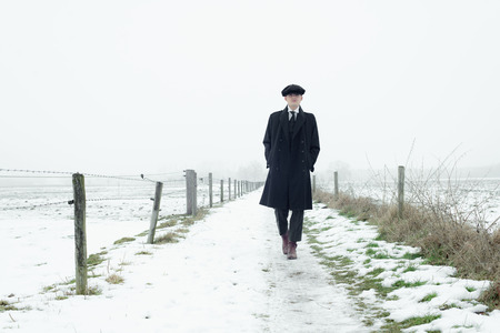 Retro 1920s english gangster with black coat and flat cap walking in winter snow landscape. Peaky blinders style.の写真素材