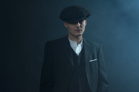 Retro 1920s english gangster wearing suit and flat cap standing in smoky room. Peaky blinders style.