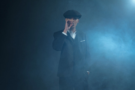 Retro 1920s english gangster wearing suit and flat cap standing in smoky room. Smoking cigarette. Peaky blinders style.の写真素材
