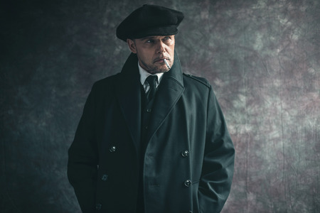 Retro 1920s english gangster smoking cigarette. Wearing black coat and flat cap.の写真素材