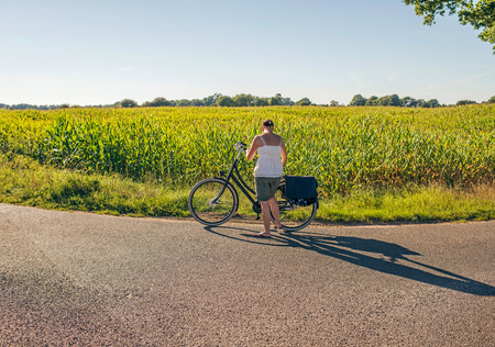 Female tourist with bicycle standing on rural road in summer.の写真素材