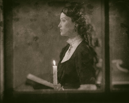 Worn wet plate photo of victorian girl reading book by candlelight behind window.