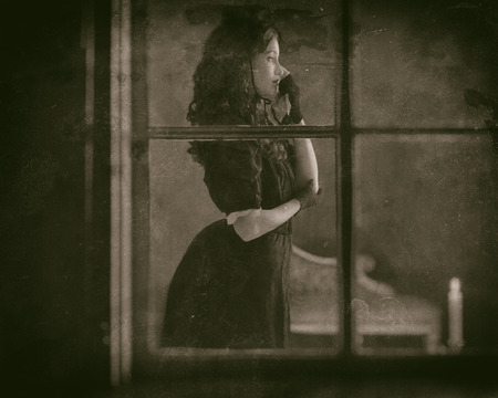 Worn wet plate photo of sad retro victorian woman in black dress wiping tear standing behind window.の写真素材
