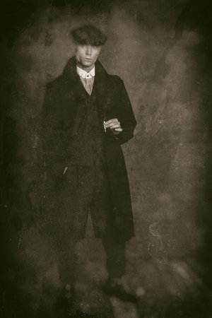 Antique wet plate photo of macho 1920s english gangster wearing black coat and flat cap holding...