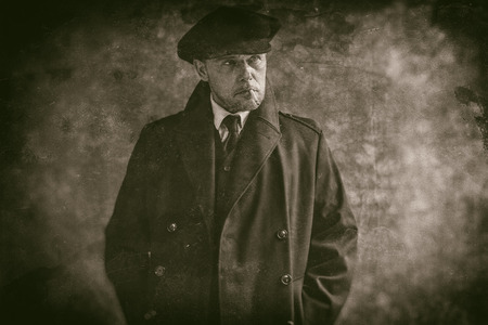 Retro 1920s english gangster smoking cigarette. Wearing black coat and flat cap.の写真素材