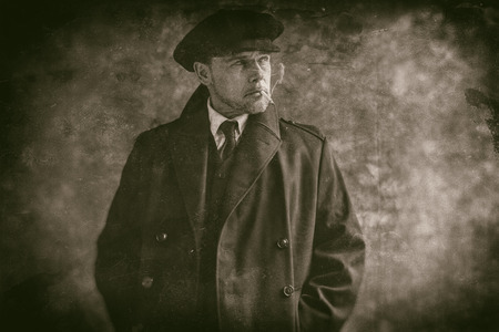 Retro 1920s english gangster smoking cigarette. Wearing black coat and flat cap.の写真素材